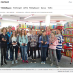 Neun Kinder und drei Betreuerinnen vor Bücherregalen in einer Bücherei. Die Kinder halten Kameras in den Händen.