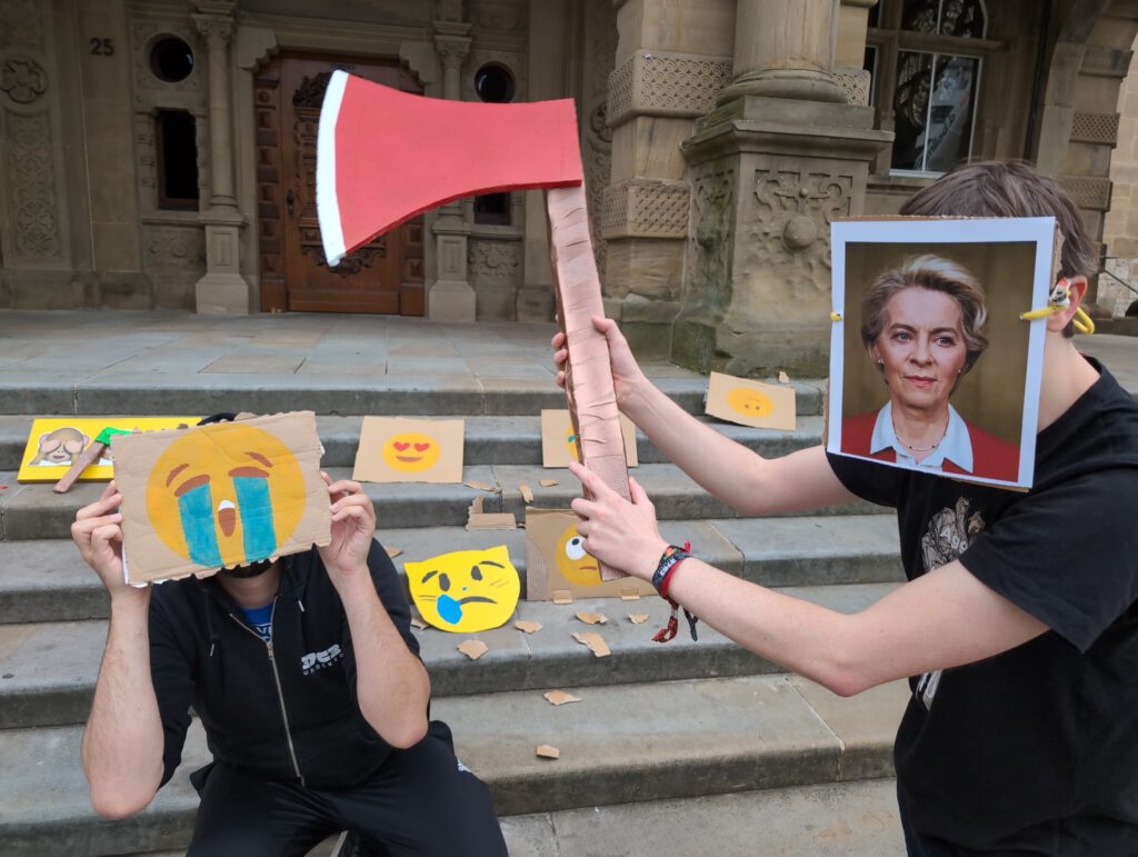 Zwei Personen vor den Treppen des Bielefelder Rathauses: Die linke Person hält sich eine Pappe vor das Gesicht auf dem ein weinendes Emoji abgebildet ist, während die rechte Person (mit einem Bild vo Ursula von der Leyen vor dem Gesicht) mit einer Akt auf sie zugeht. Das Bild stammt von der Protestaktion gegen die „Chatkontrolle“ am 17.06.24