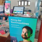 Im Vordergrund ein Plakat mit einem jungen Mann, der ein Tablet in der Hand hält und der Aufschrift "König der Suchmaschinen". Ein Infotisch mit Flyern, bunten Fruchtgummitütchen, Kühlschrankmagneten, Infohefte und Kugelschreibern. Überall ist das Logo des #DigitalCheckNRW zu sehen